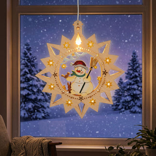 LED‑Fensterbild in Sternform aus naturfarbenem Holz mit farbig gestaltetem Schneemann-Motiv, warm beleuchtet und am Fenster aufgehängt. In der Mitte ist ein Schneemann mit roter Mütze, grünem Schal und Besen zu sehen, begleitet von kleinen Tieren und umgeben von leuchtenden Sternen. Im Hintergrund befindet sich eine verschneite Winterlandschaft; im Innenraum sind eine Pflanze, ein Sessel mit Decke und Fensterrahmen sichtbar.