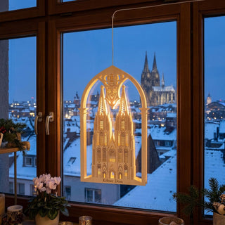 LED‑Fensterbild aus naturfarbenem Holz mit Motiv des Kölner Doms, warm beleuchtet und am Fenster aufgehängt. Das detailreiche Design zeigt die markanten Türme der Kathedrale innerhalb eines dekorativen Rundbogens mit Verzierungen. Im Hintergrund ist eine verschneite Stadtlandschaft bei Dämmerung zu sehen, in der Ferne ebenfalls der Kölner Dom; im Innenraum stehen Pflanzen, Kerzen und Dekoration auf der Fensterbank.
