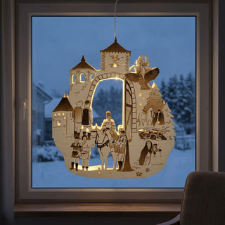 LED‑Fensterbild aus Holz mit Szene aus „Drei Haselnüsse für Aschenbrödel®“ (2025), farbig abgesetzt und warm beleuchtet. Dargestellt ist eine märchenhafte Szene mit Torbogen und Türmen, unter dem Figuren aus dem Film stehen, darunter eine Person auf einem Pferd und mehrere weitere Figuren. Rechts ist ein Baum mit Vögeln zu sehen. Das Fensterbild hängt vor einer verschneiten Winterlandschaft in der Abenddämmerung.