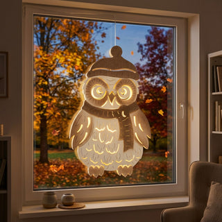 LED‑Fensterbild aus naturfarbenem Holz in Form einer Eule, warm beleuchtet und am Fenster aufgehängt. Die Eule trägt eine Mütze und einen Schal, mit leuchtenden Details in Augen und Körper. Im Hintergrund ist eine herbstliche Landschaft mit bunten Blättern und Bäumen zu sehen; im Innenraum stehen kleine Deko-Objekte auf der Fensterbank sowie ein Sessel daneben.