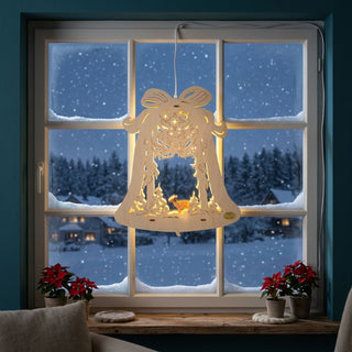 LED‑Fensterbild in Glockenform aus naturfarbenem Holz, warm beleuchtet und am Fenster aufgehängt. Im Inneren ist eine winterliche Waldszene mit handgeschnitztem Reh unter Bäumen dargestellt, darüber filigrane Stern- und Schneeflockenmuster. Im Hintergrund befindet sich eine verschneite Winterlandschaft mit Häusern; auf der Fensterbank stehen Weihnachtssterne in Töpfen.