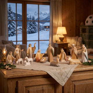 Krippenfiguren „Heilige Familie“, 15-teilig (ca. 13 cm), farbig gestaltet mit stilisierten Figuren von Maria, Josef und dem Kind, den Heiligen Drei Königen, Hirten sowie Tieren. Die Szene ist auf einem Holzmöbel arrangiert; im Hintergrund ein Fenster mit verschneiter Winterlandschaft, eine Lampe und warme Beleuchtung, die eine ruhige, festliche Atmosphäre schaffen.
