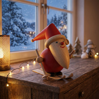 Miniatur „Weihnachtsmann auf Ski“ (ca. 8 cm), farbig gestaltet aus Holz, mit roter Kleidung, Zipfelmütze und langem Bart, auf Skiern mit Stöcken stehend. Die Figur befindet sich auf einem Holzmöbel in einer gemütlichen Wohnstube; im Hintergrund ein Fenster mit verschneiter Winterlandschaft sowie warme Lichter und winterliche Deko, die eine festliche Atmosphäre schaffen.