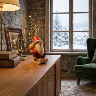 Miniatur „Weihnachtsmann auf Schlitten“ (ca. 8 cm), farbig gestaltete Holzfigur mit roter Kleidung, Zipfelmütze und langem Bart, auf einem kleinen Holzschlitten sitzend. Die Figur steht auf einem Holzmöbel in einer gemütlichen Stube; im Hintergrund ein Fenster mit verschneiter Winterlandschaft, eine Lampe, Bücher und warme Lichter, die eine festliche, winterliche Atmosphäre schaffen.