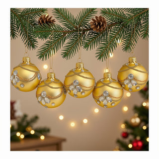 Fünf goldfarbene Christbaumkugeln aus Glas hängen an einem dekorativen Tannenzweig mit Tannenzapfen. Die Kugeln sind mit filigranen goldenen Perlenbändern, feinen Glitzerlinien und klaren, facettierten Schmucksteinen verziert. Im Hintergrund sorgen warme Lichterketten und ein festlich geschmückter Weihnachtsbaum für eine gemütliche Weihnachtsatmosphäre. Das 5-teilige Set wirkt elegant, hochwertig und besonders festlich.