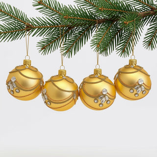 Vier goldfarbene Christbaumkugeln aus Glas hängen an den Zweigen eines Tannenbaums. Die Kugeln sind mit filigranen goldenen Perlenbändern, feinen Glitzerlinien und klaren, facettierten Schmucksteinen verziert. Durch die matte Goldoberfläche und die opulenten Dekorelemente wirkt der Christbaumschmuck besonders festlich und elegant.