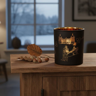 Glas-Kerzenhalter für ein Teelicht mit Motiv „Drei Haselnüsse für Aschenbrödel®“, auf einer Holzkommode vor einem Fenster platziert. Der schwarze Glaszylinder ist mit goldfarbenen Illustrationen verziert, die ein Schloss sowie Figuren und Szenen aus dem Märchen zeigen. Im Inneren sorgt ein Teelicht für warmes Licht, das die Motive zum Leuchten bringt; daneben liegen herbstliche Blätter und Haselnüsse, im Hintergrund eine winterliche Landschaft.