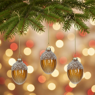 Christbaumschmuck „Drei Haselnüsse für Aschenbrödel®“ aus Glas im 3er-Set (6 cm), dekorative Glaskugeln in Haselnussform mit glitzernder Kappe, am Tannenzweig aufgehängt, vor warm leuchtendem Weihnachtslichter-Hintergrund.