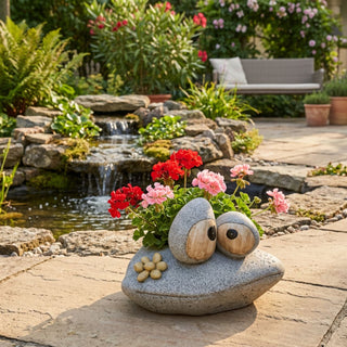 Pflanzgefäß in Froschform (ca. 26 cm) aus Polyresin im Stein-Design, mit großen Augen und bepflanzt mit roten und rosa Blüten. Steht auf einer Terrasse vor einem kleinen Gartenteich mit Steinen und Pflanzen.
