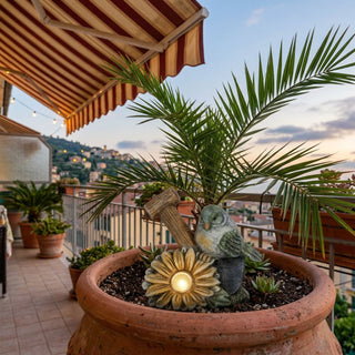 Vogel aus Keramik mit Solar-LED (ca. 13 cm), sitzt neben einer Blüte mit leuchtendem Licht. Platziert in einem Blumentopf auf einem Balkon mit Blick auf eine Stadt und Pflanzen im Hintergrund.