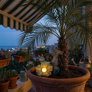 Schnecke aus Keramik mit Solar-LED (ca. 13 cm), sitzt auf einer Blüte mit leuchtendem Licht. Platziert in einem Blumentopf auf einem Balkon bei Abenddämmerung, umgeben von Pflanzen und mit Blick auf eine Stadt.