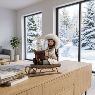 Winterkind mit Schlitten und Schneekugel (ca. 11 cm), farbig gestaltete Figur mit winterlicher Kleidung und Fellmütze, sitzend auf einem Schlitten und eine Schneekugel mit winterlichem Motiv haltend. Die Szene steht auf einem modernen Holzmöbel; im Hintergrund große Fenster mit verschneiter Winterlandschaft sowie dezente Wohnraumdekoration, die eine ruhige, winterliche Atmosphäre schaffen.