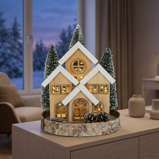 LED Dekoration „Waldhaus“ (24 cm) in naturfarbener Holzoptik auf Holzsockel, warmweiß beleuchtet, detailreiches Häuschen mit verschneiten Tannenbäumen und winterlichen Details, als stimmungsvolle Tischdekoration im gemütlichen Wohnambiente.
