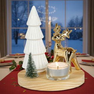 Kerzenhalter auf rundem Holzsockel für ein Teelicht mit dekorativem Motiv aus Tannenbaum und Hirsch. Neben dem eingesetzten Glas-Teelichthalter mit brennender Kerze stehen ein weißer stilisierter Baum sowie ein goldfarbener Hirsch und ein kleines grünes Bäumchen. Das Arrangement ist auf einem festlich gedeckten Tisch mit roter Tischdecke platziert; im Hintergrund sind Kerzen, Geschirr und ein Fenster mit verschneiter Winterlandschaft zu sehen.