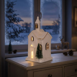 LED Keramikkirche (15 cm) für Batteriebetrieb, warmweiß beleuchtet, mit Holzfenstern und filigranen Details, dekoriert mit kleinem Tannenbaum vor der Kirche, als stimmungsvolle Winter‑ und Tischdekoration vor verschneiter Fensterlandschaft.