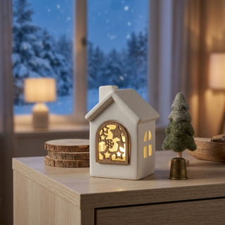 LED Keramikhaus (10 cm) mit Holzfenster, batteriebetrieben, warmweiß beleuchtet, mit dekorativem Stern‑ und Schneeflockenmotiv im Fenster, als stimmungsvolle Tisch- oder Fensterbankdekoration vor winterlicher Kulisse.