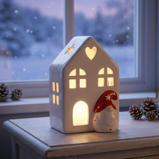 LED Keramikhaus (11 cm) mit Wichtelfigur, batteriebetrieben, warmweiß beleuchtet, weißes Haus mit ausgestanzten Stern-, Herz- und Fensterdetails, als stimmungsvolle Fensterbank‑ oder Tischdekoration vor winterlicher Kulisse.