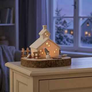 LED Keramikhaus (16 cm) auf Holzsockel, warmweiß beleuchtet, detailreiche Szene mit Haus, Zaun und kleiner Figur auf naturbelassener Basis, als stimmungsvolle Tischdekoration vor winterlicher Fensterkulisse.