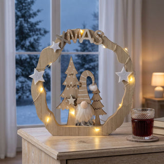 LED Dekoration „Xmas mit Wichtel“ (18 cm) in naturfarbener Holzoptik, warmweiß beleuchtet, mit integriertem Schriftzug, Sternen und Wichtelfigur, als stimmungsvolle Tischdekoration vor winterlicher Fensterkulisse.