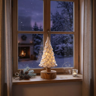 Beleuchteter LED-Keramikbaum (18 cm) auf rundem Holzsockel, dekorativ auf einer Fensterbank platziert; der Baum strahlt warmes Licht aus. Im Hintergrund ein Fenster mit winterlicher Schneelandschaft, davor weihnachtliche Dekoelemente wie Tannenzapfen, Zweige, ein Drahtornament mit Schneeflocke und ein kleines Teelichtglas.