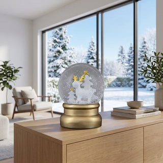 Schneekugel „Elch“ (ca. 12 cm) mit goldfarbenem Sockel und winterlichem Motiv im Inneren: ein stilisierter Elch mit goldenem Geweih neben Tannenbäumen. Die Schneekugel steht auf einem modernen Holzmöbel vor großen Fenstern mit verschneiter Winterlandschaft; dezente Wohnraumdekoration sorgt für eine ruhige, festliche Atmosphäre.