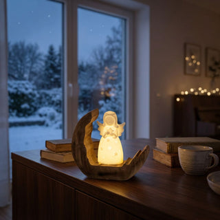 LED-Dekoration „Engel auf Holz“ (15 cm) mit leuchtender Engelsfigur in einer geschwungenen Holzform, warm beleuchtet auf einer Kommode vor winterlicher Fensterkulisse platziert.