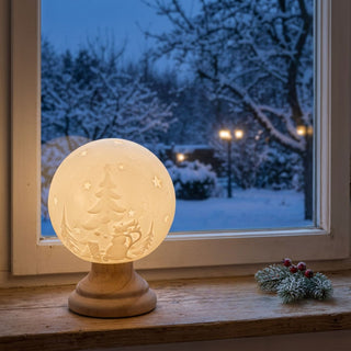 Leuchtende LED-Keramikkugel (14 cm) auf Holzsockel mit winterlichem Relief aus Tannenbaum, Sternen und Schneemann, warm beleuchtet auf einer Fensterbank platziert. Im Hintergrund ein verschneiter Garten in der Dämmerung, daneben ein kleiner Tannenzweig mit roten Beeren als dekoratives Element.