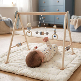 "Baby spielt auf dem Rücken mit Spielbogen 'Teddy' in Natur-Blau auf weicher Krabbeldecke – fröhlicher Entdeckungsmoment im hellen Kinderzimmer"