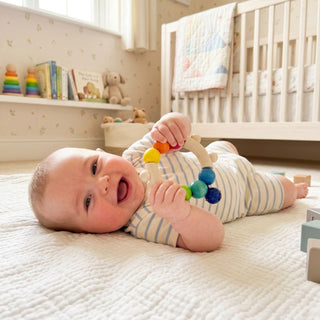 Baby liegt auf einer weichen Decke im Kinderzimmer und hält eine runde Holzrassel mit bunten Perlen in Regenbogenfarben (Ø ca. 10 cm) in den Händen; im Hintergrund stehen ein Kinderbett, Bücher und Spielzeugregal.