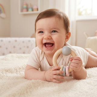 Baby liegt auf einer weichen Decke im Kinderzimmer und hält eine runde Holzrassel in Natur- und Rosatönen (Ø ca. 11 cm) mit kleinen Perlen und Ringen in der Hand; im Hintergrund sind ein Kinderbett und ein Regal mit Spielzeug zu sehen.