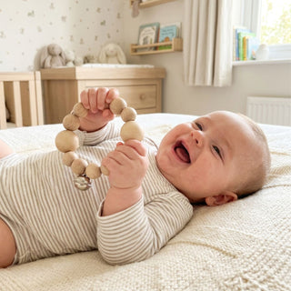 Baby liegt auf einer weichen Decke im Kinderzimmer und hält einen runden Greifling aus Naturholz mit kleinen Perlen und einer integrierten Glocke (Ø ca. 9 cm) in den Händen; im Hintergrund sind Wickelkommode, Bücher und Stofftiere zu sehen.