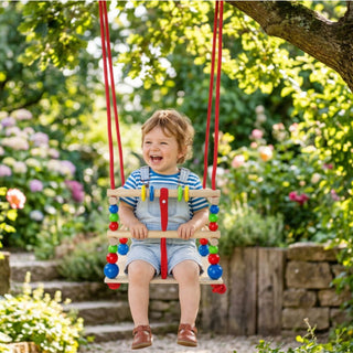 Gitterschaukel in bunten Farben mit rot-weiß gestreiftem Mittelteil und farbigen Holzkugeln in Rot, Blau, Gelb und Grün – fröhliches Kind schaukelt im sonnigen Garten zwischen blühenden Hortensien und saftig grünem Laub – unbeschwerte Sommeratmosphäre im natürlichen Outdoor-Setting