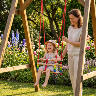 Gitterschaukel de Luxe 'farbig' mit rot-weiß gestreiftem Mittelteil und bunten Holzkugeln in Rot, Blau, Gelb und Grün – fröhliches Kind schaukelt mit Mama im sonnigen Garten zwischen blühenden Rittersporn und Rosen – unbeschwerte Sommeratmosphäre im natürlichen Outdoor-Setting mit Holzschaukelgestell