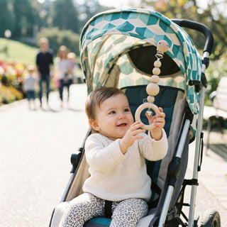 Kleinkind sitzt im Kinderwagen im Freien und greift nach einem natürlichen Kinderwagenketten-Anhänger aus Holz mit Perlen, Ring und kleiner Glocke (ca. 20 cm lang), der am Verdeck befestigt ist; im Hintergrund ein sonniger Parkweg mit Bäumen und Spaziergängern.