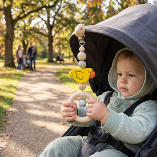 Kleinkind sitzt im Kinderwagen im Freien und hält einen Holzring, an dem eine Kinderwagenkette mit buntem Küken-Anhänger, Perlen und kleiner Glocke (ca. 20 cm lang) befestigt ist; im Hintergrund sind Bäume und ein parkähnlicher Weg zu sehen.