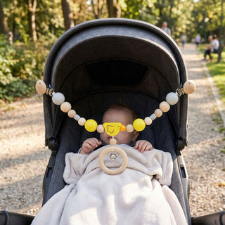 Baby liegt im Kinderwagen im Freien und blickt auf eine befestigte Kinderwagenkette mit buntem Küken-Motiv, Holzperlen, Ring und integrierter Glocke; die Kette ist mit zwei Clips am Verdeck befestigt und ca. 52 cm lang, im Hintergrund ein parkähnlicher Weg mit Bäumen.
