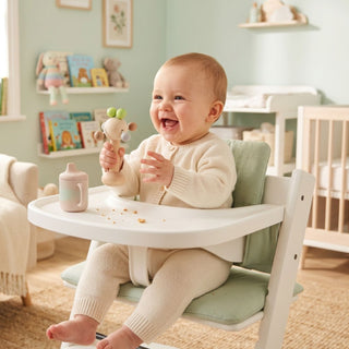 Kind sitzt im Hochstuhl im Kinderzimmer und hält eine Holzrassel „Klapperbäumchen“ in Natur- und Apfelgrüntönen (ca. 13 cm) mit beweglichen Elementen in der Hand; auf dem Tisch liegen Essenskrümel, im Hintergrund sind Regale, Bücher, Kuscheltiere und ein Babybett zu sehen.