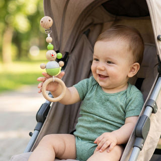 Kind sitzt im Kinderwagen im Freien und greift nach einem bunten Greifling mit Maus-Motiv, befestigt an einem Clip; der Anhänger besteht aus Holzperlen und einem Ring (ca. 20 cm lang), im Hintergrund ein von Bäumen gesäumter Weg.