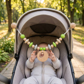Baby sitzt im Kinderwagen im Freien und betrachtet eine farbige Kinderwagenkette mit Kreis‑Element, Holzperlen und Ring, die mit zwei Clips am Verdeck befestigt ist (ca. 47 cm lang); im Hintergrund ein sonniger Parkweg mit Bäumen.