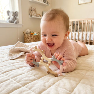 Baby liegt auf einer weichen Decke im Kinderzimmer und hält eine Motorikrassel in Form einer Acht aus Holz in Natur- und Rosatönen mit beweglichen Perlen in den Händen; im Hintergrund sind ein Kinderbett, Regale und Stofftiere zu sehen.