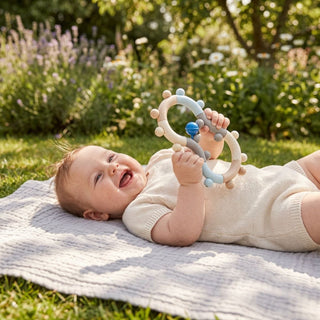 Baby liegt auf einer Decke im Freien und hält eine Motorikrassel in Form einer Acht aus Holz in Natur- und Blautönen mit beweglichen Perlen und Ringen in den Händen; im Hintergrund eine sonnige Gartenlandschaft mit Blumen und Bäumen.