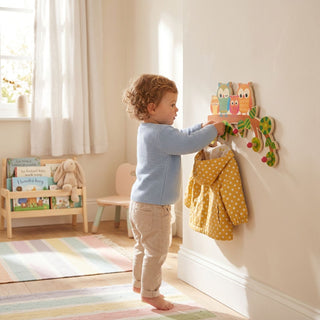 Garderobe „Eulen“ mit 5 Haken, bunt gestaltet mit mehreren Eulenmotiven. Ein Kind hängt eine gelbe Jacke an die Haken in einem hellen Kinderzimmer; im Hintergrund sind ein kleines Bücherregal, Spielzeug und ein Fenster zu sehen.