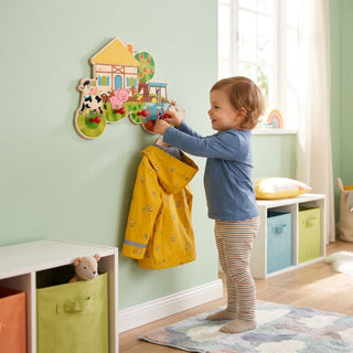 Garderobe „Bauernhof“ mit 5 Haken, bunt gestaltet mit Motiven wie Kuh, Schwein und Traktor. Ein Kind hängt eine gelbe Jacke an die Haken in einem hellen Kinderzimmer; im Hintergrund sind Aufbewahrungsboxen, Spielzeug und ein Fenster zu sehen.