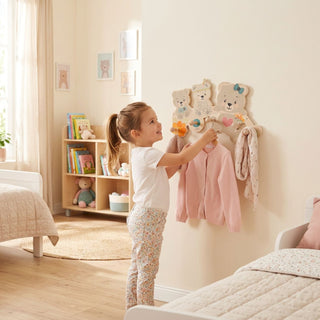 Garderobe „Bärchen“ mit 5 Haken, farbig gestaltet mit niedlichen Bärenmotiven. Ein Kind hängt Kleidung an die Haken in einem hellen Kinderzimmer; im Hintergrund stehen ein Regal mit Büchern und Spielzeug sowie ein Bett.