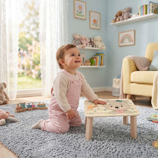 Fußbank 'Bärchen' aus Holz in bunten Farben mit freundlichem Bärchen-Gesicht und Regenbogen-Motiv auf der Sitzfläche – Kleinkind spielt im hellen, freundlichen Kinderzimmer mit sanft blauer Wand, weißen Holzregalen mit Kuscheltieren und Kinderbüchern, gelbem Sessel und flauschigem hellgrauem Teppich – unbeschwerte Spielatmosphäre im natürlichen Wohnambiente