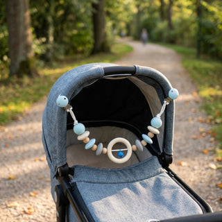 Leerer Kinderwagen steht im Freien auf einem Weg; daran ist eine Kinderwagenkette in Natur- und Blautönen mit Holzperlen, Ring und kleiner Glocke befestigt, die mit zwei Clips am Verdeck fixiert ist, im Hintergrund ein von Bäumen gesäumter Parkweg.