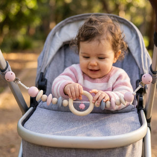 Kleinkind sitzt im Kinderwagen im Freien und greift nach einer Kinderwagenkette in Natur- und Rosatönen mit Holzperlen, Ring und kleiner Glocke; die Kette ist mit zwei Clips am Verdeck befestigt, im Hintergrund ein sonniger Parkweg mit Bäumen.