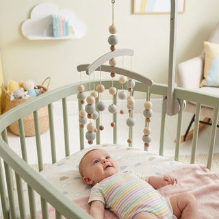 Baby liegt im Kinderbett und blickt nach oben auf ein hängendes Kugel-Mobile aus Holz in Natur- und Grautönen (ca. 52 cm) mit unterschiedlich großen Perlen; das Mobile ist an einem Gestell befestigt, im Hintergrund ein liebevoll eingerichtetes Kinderzimmer mit Regal, Körben und Sitzmöbel.