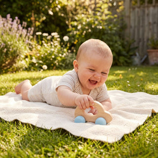 Baby liegt auf einer Decke im Garten und schiebt eine Holz-Schiebefigur „Rolli Auto“ in Natur- und Blautönen mit kleinen Rädern über den Boden; im Hintergrund eine sonnige Gartenlandschaft mit Blumen und Grünflächen.