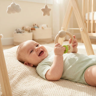 Baby liegt auf einer weichen Decke im hellen Kinderzimmer und hält eine runde Holzrassel in Natur- und Apfelgrüntönen (Ø ca. 9 cm) mit beweglichen Ringen in den Händen; im Hintergrund sind ein Spielbogen sowie Körbe und gehäkelte Dekoelemente zu sehen.
